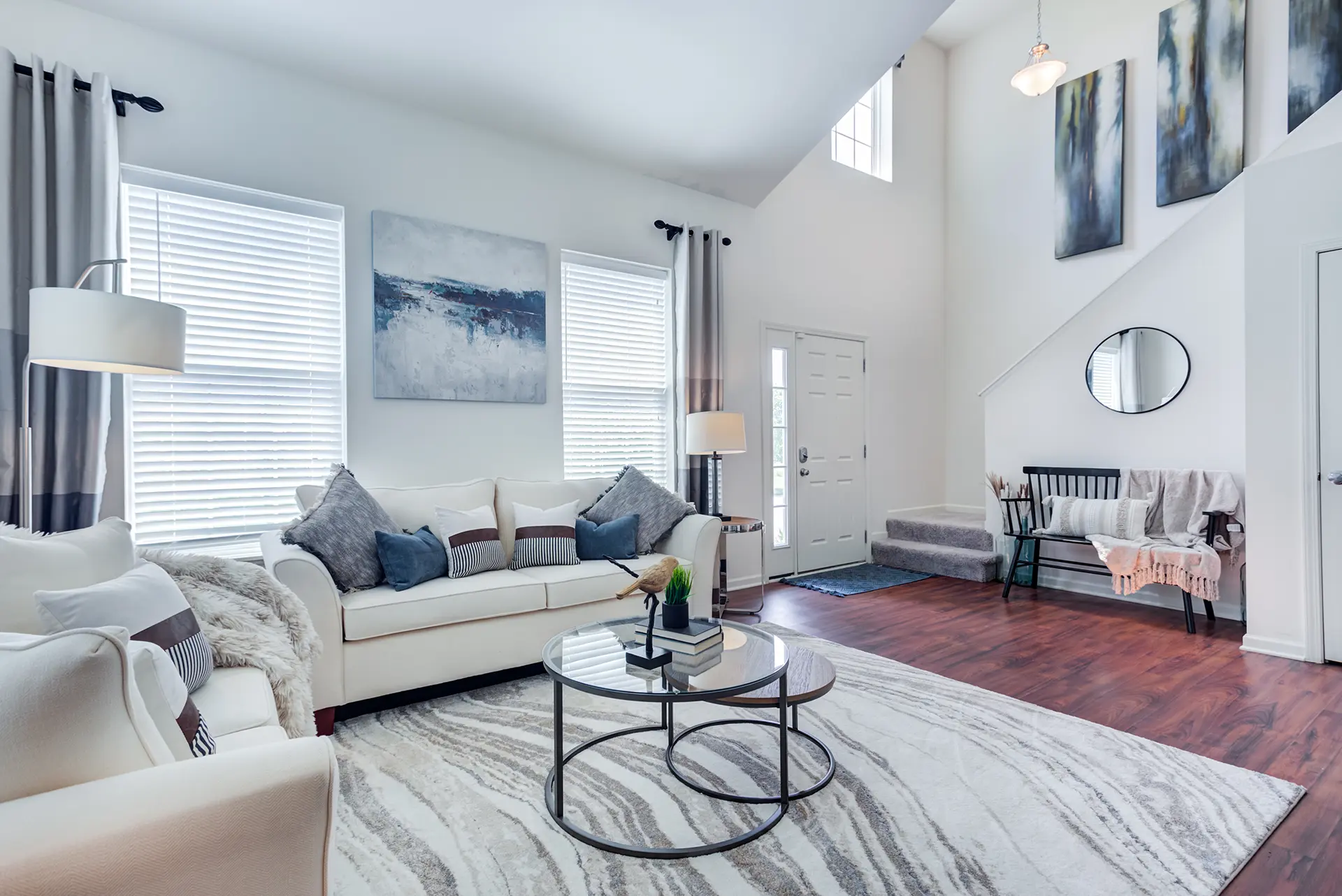 Bright, modern living room with white walls, large windows, high ceilings, two white sofas, glass table, wood-look floors, and abstract art.