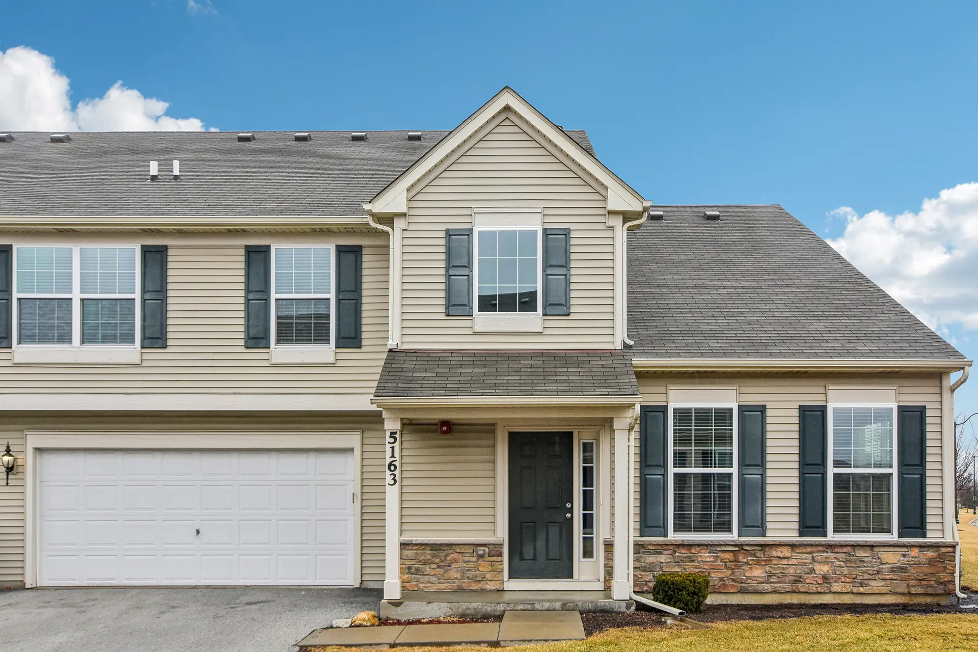 A beige two-story house with blue shutters, double garage, stone accent, wood-look porch, number 5163, and small partly brown lawn.