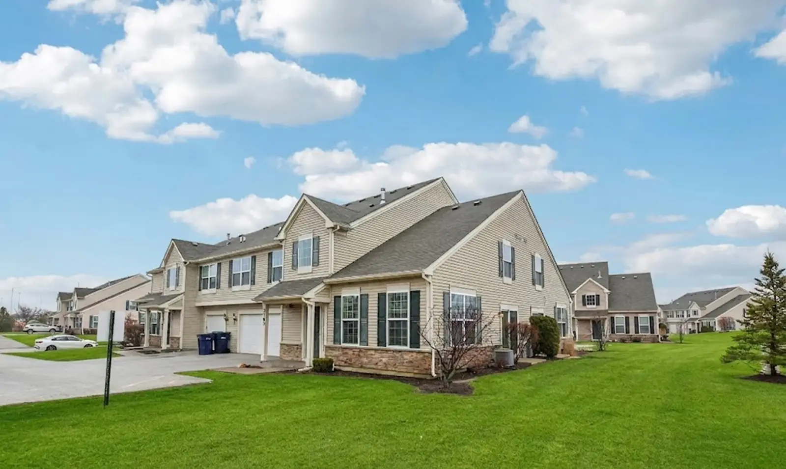 Two beige, two-story townhouses with wood-look attached garages sit on neat green lawns under a blue sky with scattered clouds.
